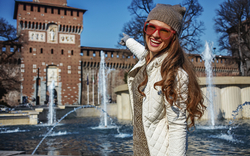 Eine Frau mit langen Haaren, warmer Kleidung und einer Sonnenbrille steht lächelnd vor einem Brunnen mit Wasserfontänen. Im Hintergrund ist ein historisches Gebäude aus Backstein zu sehen.