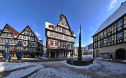 Ein Platz im Winter mit Schnee bedeckt, umgeben von Fachwerkhäusern und einem Brunnen in der Mitte.