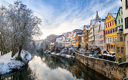 Verschneite Stadtansicht an einem Fluss mit bunten Häusern und Bäumen im Winter.